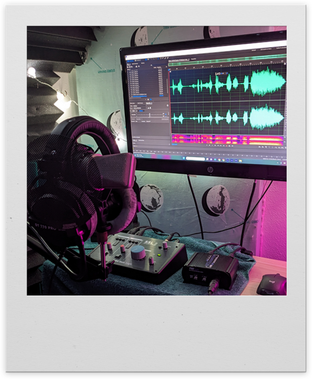 A recording setup. Headphones sit on a microphone stand that holds a Neumann TLM103 microphone. A small audio interface sits on a raised desk. Adobe Audition is on a monitor showing a recently recorded waveform.
